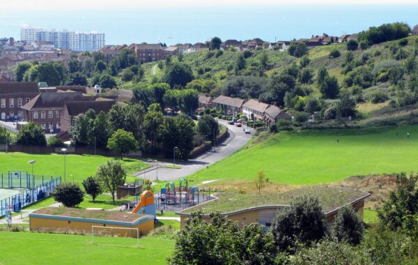 A view of Whitehawk Hill in Brighton. Green space and aplayground area are visible in the middle distance, with housing and then the sea in the background.
