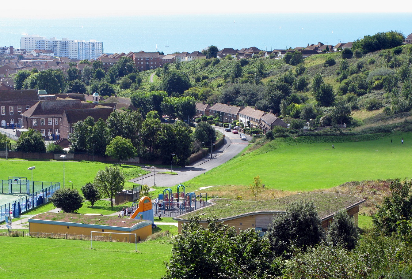 A view of Whitehawk Hill in Brighton. Green space and aplayground area are visible in the middle distance, with housing and then the sea in the background.