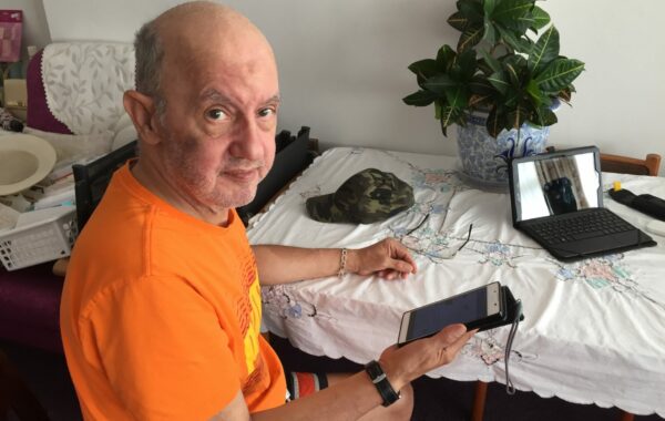 Giorgio sits at a table, facing the camera and holding his mobile phone. He is wearing a bright orange t-shirt. On the table is a pot plant and a laptop.